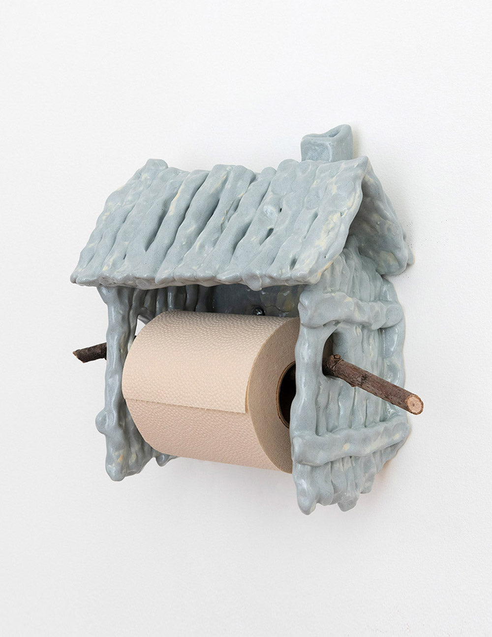 Three-quarter view of ceramic house-shaped toilet paper holder showing twig spindle and textured glazed roof on white wall